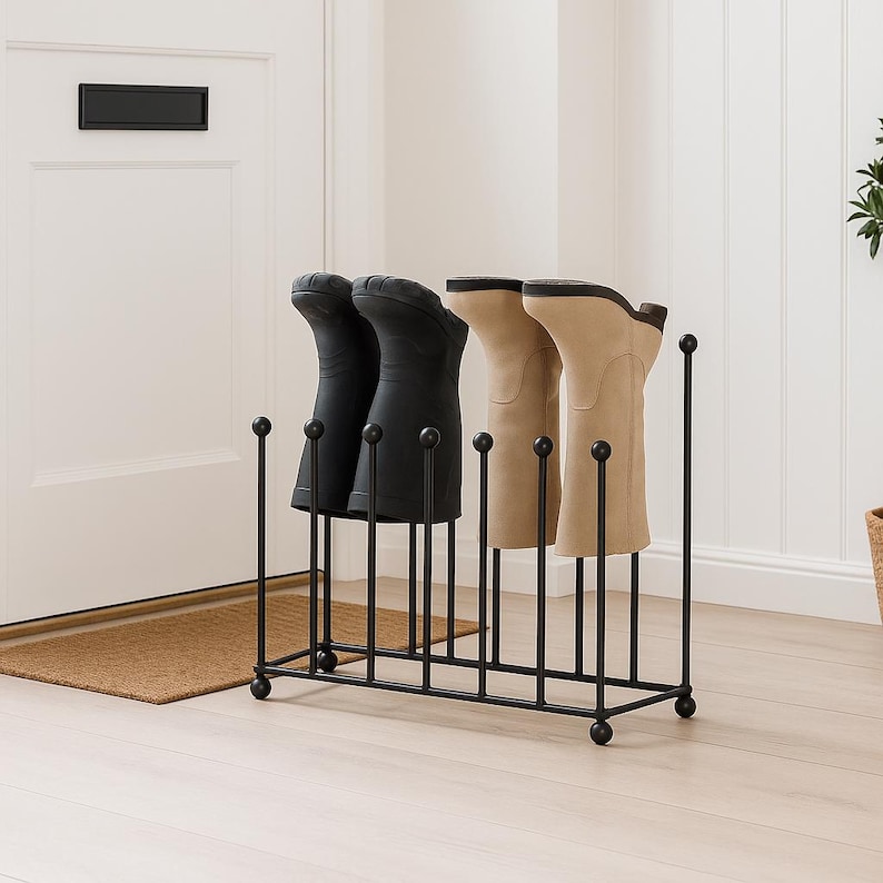 May include: A black metal boot rack holding two pairs of boots. The boots are black and tan. The rack has a rectangular base with vertical supports and decorative ball finials. The rack is on a light wood floor.