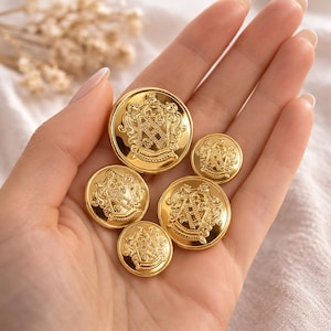 May include: A collection of five gold-colored buttons with ornate crest designs. The buttons vary in size and are arranged in a hand. The crests feature intricate details and are set against a shiny, reflective surface.