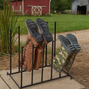 May include: A black metal boot rack holding two pairs of boots. One pair is brown leather, the other is camouflage. The rack is on a concrete slab, with a red barn in the background.