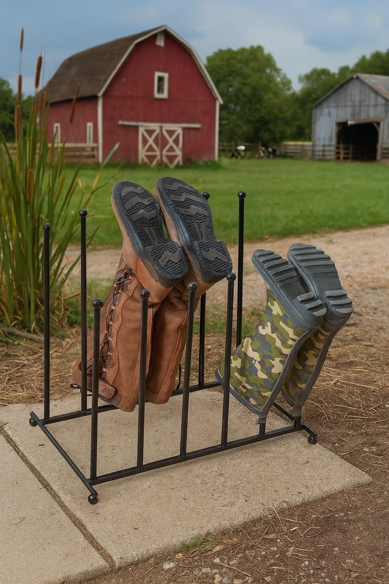 May include: A black metal boot rack holding two pairs of boots. One pair is brown leather, the other is camouflage. The rack is on a concrete slab, with a red barn in the background.