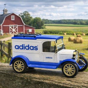 May include: A vintage-style toy truck, white with blue accents, features the Adidas logo on its side. The truck has gold-colored wheels and is set against a rural backdrop with a red barn and hay bales.