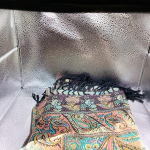 May include: A folded scarf with a paisley pattern in pink, teal, purple, and gold. It has a black fringe. The scarf is inside a silver box. The design is intricate and colourful.