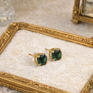 May include: Gold-coloured stud earrings featuring square, emerald-green gemstones. The earrings are presented on a gold-framed surface with a delicate, lacy pattern. The image also includes a decorative frame and a glass container.