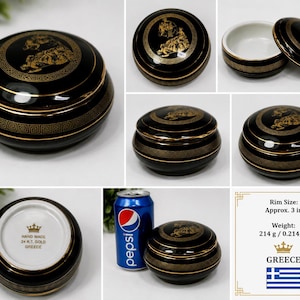 May include: A black and gold ceramic trinket box with a lid. The box has a Greek key design and a gold illustration on the lid. The rim size is approximately 7.6 cm. The box is labeled "HAND MADE 24 KT. GOLD GREECE".
