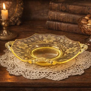 May include: A yellow glass serving dish with floral etchings, on a white lace doily. The dish has handles and is surrounded by antique items, including a candle, books, and a jewelry box, creating a nostalgic aesthetic.
