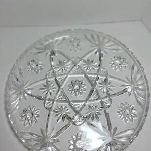 May include: A clear, round glass serving plate with a starburst and floral design. The plate features a central star pattern surrounded by radiating lines and smaller floral accents. The edges are scalloped, adding a decorative touch to this elegant piece.