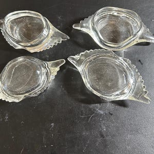 May include: Four clear glass ashtrays with a distinctive design. Each ashtray features a circular bowl shape, a pointed end, and a decorative, scalloped edge. The ashtrays are arranged on a dark surface.