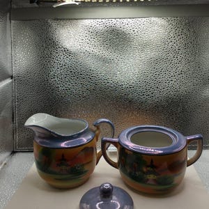 May include: A vintage ceramic creamer and sugar bowl set with a lid. The set features a landscape design with a pagoda, trees, and mountains. The rims and lid have a blue iridescent finish, and the bodies are a mix of orange, yellow, and green.