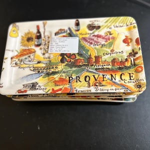 May include: A stack of rectangular ceramic trays with a colorful map of Provence, France. The trays feature illustrations of sunflowers, cheese, and wine bottles. The word "PROVENCE" is prominently displayed.