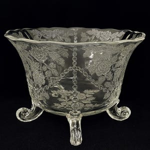 Puede incluir: Cuenco de vidrio transparente con pie y diseños florales y de hojas grabados. El cuenco tiene un borde festoneado y tres patas ornamentadas. El diseño grabado incluye rosas y un patrón en forma de cadena. El cuenco es probablemente para fines decorativos o de servicio.