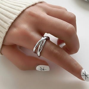 Op de afbeelding: Een zilverkleurige ring met een drievoudig bandontwerp. De ring wordt aan een vinger gedragen, met een witte mouw op de achtergrond. De banden van de ring kruisen elkaar, wat een moderne uitstraling creëert. Een witte marmeren steen staat op de achtergrond.