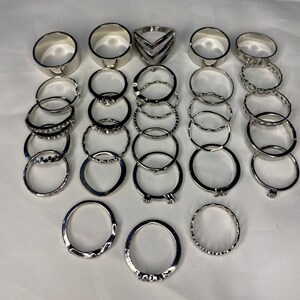 May include: A set of 20 silver-toned rings, featuring a variety of styles, including plain bands, textured bands, and geometric designs.