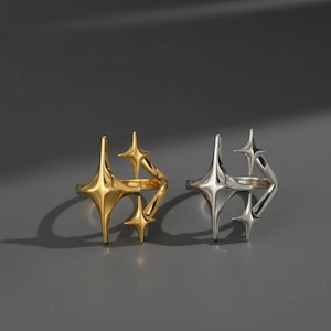 Könnte beinhalten: Zwei Ringe, einer goldfarben und einer silberfarben, jeweils mit einem Sternenmuster. Die Ringe haben eine moderne, geometrische Ästhetik mit mehreren Sternformen, die um das Band angeordnet sind. Der Hintergrund ist ein neutrales Grau.