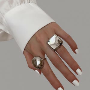 Puede incluir: Dos anillos plateados se muestran en una mano con esmalte de uñas blanco. Uno tiene un diseño grande, redondeado y liso, mientras que el otro presenta una forma geométrica cuadrada. Los anillos reflejan la luz, creando una apariencia brillante.