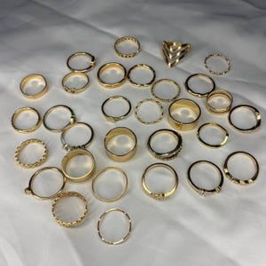 May include: A set of 25 gold-toned rings, featuring a variety of styles, including simple bands, textured bands, and rings with geometric designs.