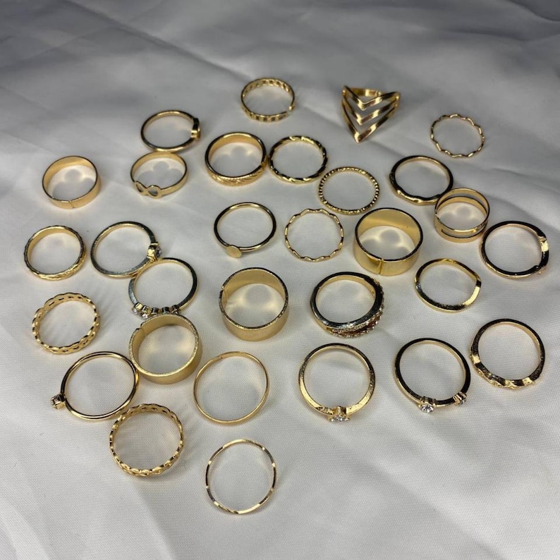 May include: A set of 25 gold-toned rings, featuring a variety of styles, including simple bands, textured bands, and rings with geometric designs.