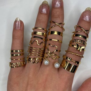 May include: A set of gold-toned rings featuring various designs, including infinity, hearts, butterflies, and wavy lines. The rings are stacked on a hand, with two pearl-like accents on the middle finger.