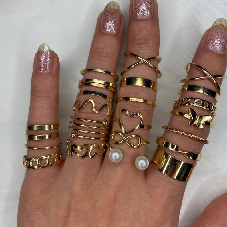 May include: A set of gold-toned rings featuring various designs, including infinity, hearts, butterflies, and wavy lines. The rings are stacked on a hand, with two pearl-like accents on the middle finger.
