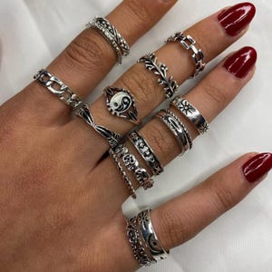 May include: A selection of silver rings on fingers. The rings have diverse designs, such as chain links, leaves, skulls, and a yin-yang symbol. The rings are worn on multiple fingers, highlighting their varied styles and textures.