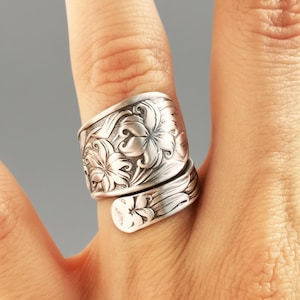 May include: A silver-toned spoon ring with a floral design. The ring wraps around the finger, featuring detailed carvings of flowers and leaves. The ring is a unique piece of jewelry.