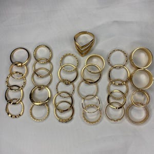 May include: A set of 20 gold-toned rings, featuring a variety of styles and designs, including plain bands, textured bands, and rings with small accents. The rings are arranged in rows on a white background.