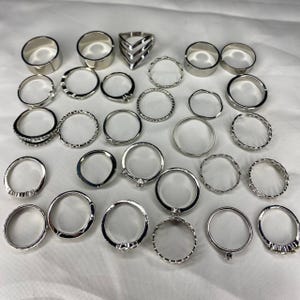 May include: A set of 24 silver-toned rings, featuring a variety of styles, including plain bands, textured bands, and rings with small accents. The rings are arranged on a white surface.