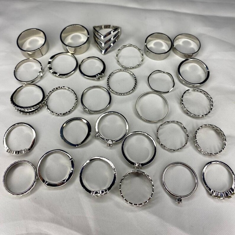 May include: A set of 24 silver-toned rings, featuring a variety of styles, including plain bands, textured bands, and rings with small accents. The rings are arranged on a white surface.