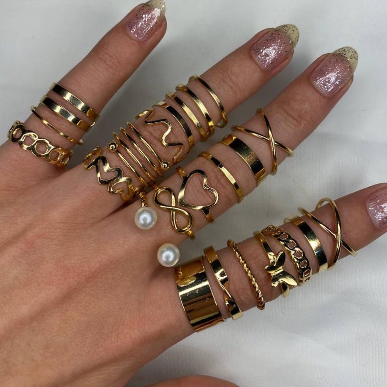 May include: A set of 18 gold-toned rings featuring various designs, including infinity symbols, hearts, and simple bands. The rings are worn on multiple fingers, creating a layered look.