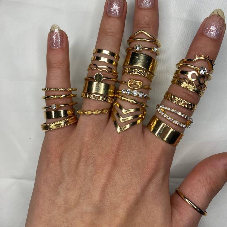 May include: A set of 18 gold-toned rings with various designs, including a wave, a circle, a knot, a chevron, and a ring with small clear stones. The rings are stacked on a hand.