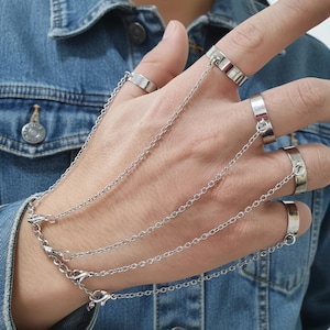 Peut inclure: Bracelet chaîne de main argenté avec plusieurs bagues reliées par de fines chaînes. Les bagues sont portées aux doigts et attachées à un fermoir de bracelet. Le bijou est en métal brillant.