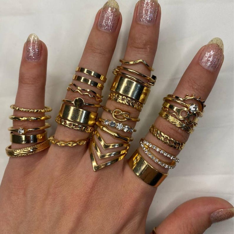 May include: A set of gold-toned rings with various designs, including a chain, a circle with a small stone, a chevron, and a simple band. The rings are stacked on a hand, creating a layered look.
