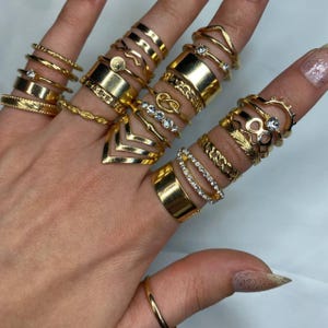 May include: A set of 18 gold-toned rings with various designs, including infinity, knot, and geometric shapes. Some rings feature small clear rhinestones.