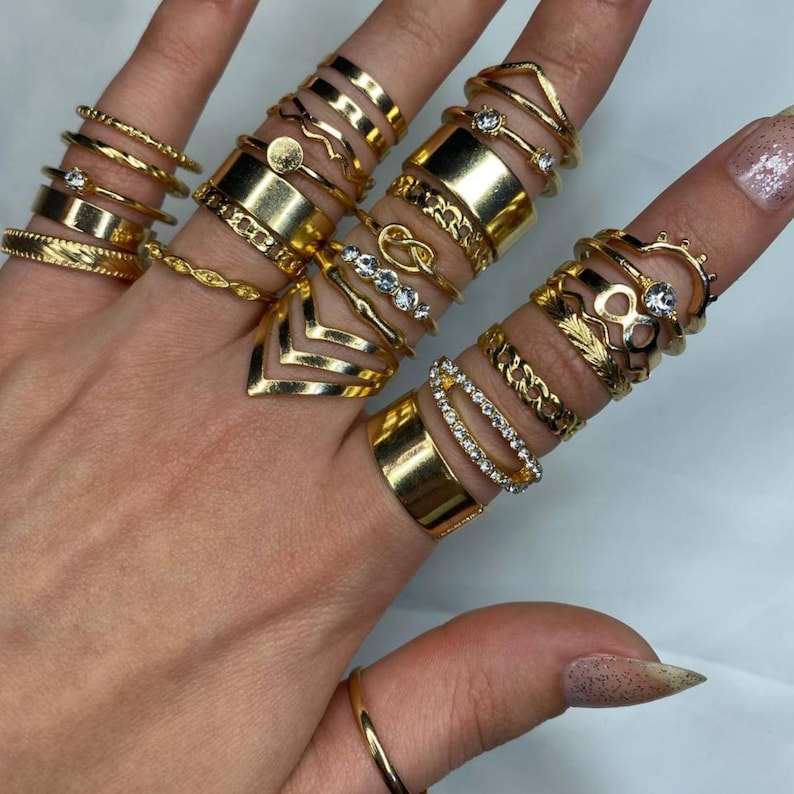 May include: A set of 18 gold-toned rings with various designs, including infinity, knot, and geometric shapes. Some rings feature small clear rhinestones.