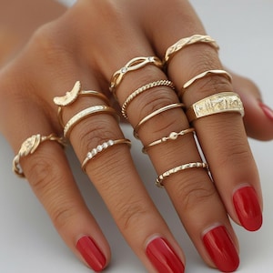 Op de afbeelding: Een verzameling goudkleurige ringen in verschillende ontwerpen, waaronder banden met getextureerde details, kleine edelstenen en een halve maanvorm. De ringen worden op meerdere vingers gedragen en tonen hun verschillende stijlen.