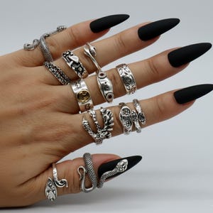 May include: A collection of silver rings in various designs, including snake, floral, and geometric patterns. The rings are displayed on a hand with long, black, pointed fingernails. The rings vary in size and style, showcasing intricate details.