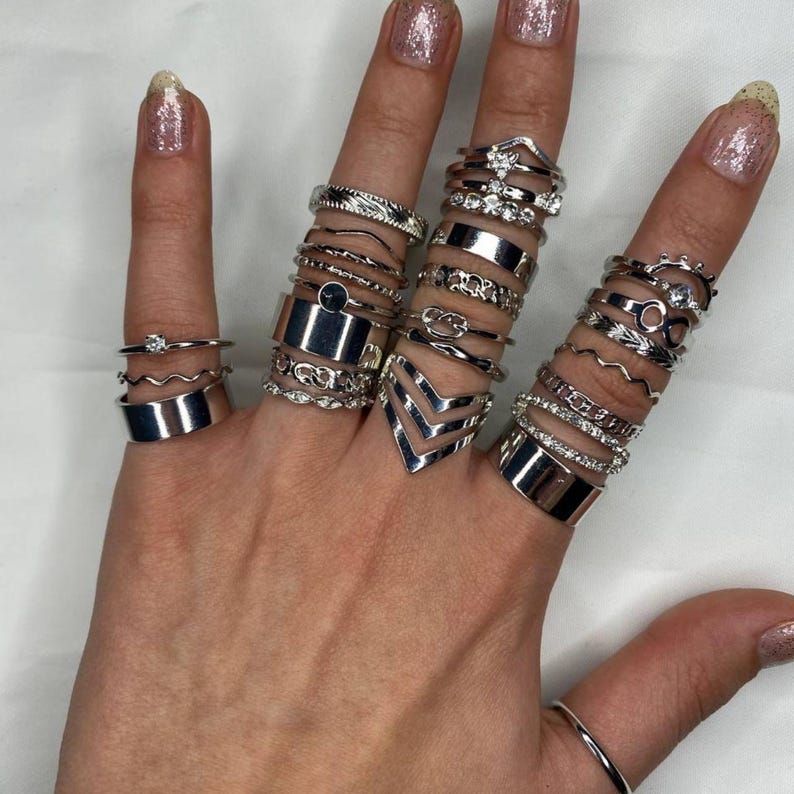 May include: A hand wearing a stack of silver rings. The rings are of various styles, including simple bands, geometric shapes, and designs with small stones.