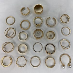 May include: A set of 24 gold-toned rings, featuring a variety of styles, including simple bands, twisted designs, and open rings with pearl accents.