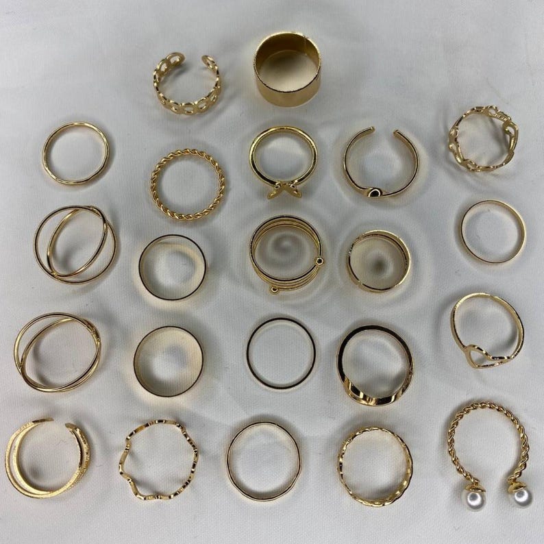 May include: A set of 24 gold-toned rings, featuring a variety of styles, including simple bands, twisted designs, and open rings with pearl accents.