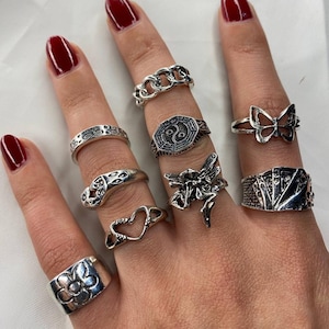 May include: A hand model wearing a set of nine silver rings. The rings feature various designs, including a heart, a flower, a butterfly, a yin yang symbol, a snake, a fairy, and a playing card design.