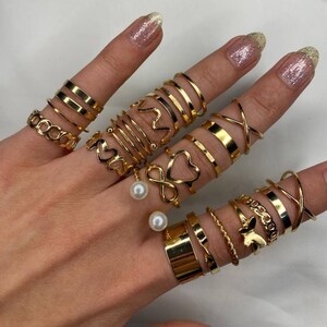 May include: A set of gold-toned rings featuring various designs, including hearts, infinity symbols, and simple bands. The rings are worn on multiple fingers, creating a layered look. Two pearl rings are also included.
