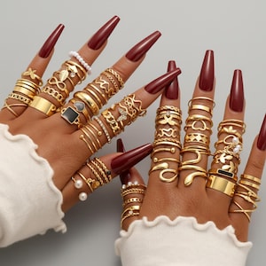 Könnte beinhalten: Eine Sammlung goldfarbener Ringe in verschiedenen Designs schmückt Finger mit langen, burgunderfarbenen Nägeln. Die Ringe umfassen Bänder, Perlen und dekorative Stile, darunter Schmetterlinge, Herzen und geometrische Formen. Die Hand trägt einen weißen Pullover.