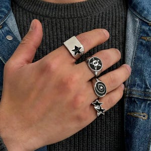 Op de afbeelding: Een hand toont verschillende zilveren ringen met diverse ontwerpen. Er zijn sterren, cirkels en spiralen te zien. De ringen worden gedragen tegen een denim jas.