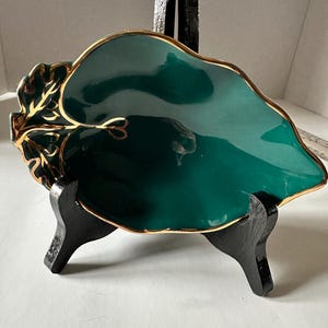 Peut inclure: Un plat décoratif en forme de feuille, vert émeraude profond avec une bordure dorée. Le plat est présenté sur un présentoir noir. Le plat a une finition brillante et est probablement en céramique ou en porcelaine. La bordure dorée ajoute une touche d'élégance.