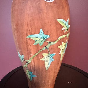 May include: A tall, terracotta-coloured vase with a unique shape and a white rim. The vase is adorned with a painted branch featuring green and blue leaves. The vase is sitting on a dark surface.