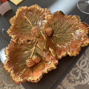 May include: A decorative, leaf-shaped serving dish with a gold finish. The dish features intricate veining and is accented with gold trim. It has a handle and is adorned with three small, textured, gold-coloured decorative elements.