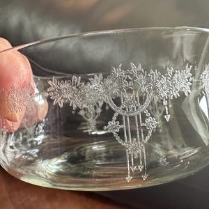 May include: A clear glass bowl featuring a delicate, etched silver-toned floral pattern that wraps around the exterior. The design includes intricate blossoms, hanging geometric pendants, and fine lines, creating a detailed decorative motif.
