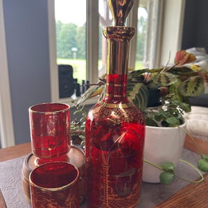 May include: A tall red glass decanter with a gold stopper and two matching red glass tumblers. Each piece features intricate gold filigree patterns and detailed illustrations of historical buildings and figures, accented with gold rims.