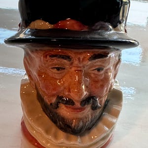 May include: A ceramic mug in the shape of a man's head, with a black hat, a tan face with a mustache and beard, and a white ruffled collar. The mug has a glossy finish.