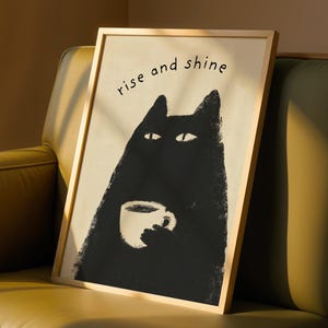 May include: A framed art print featuring a black cat holding a coffee cup, with the words "rise and shine" in black script. The artwork has a minimalist style and is set against a cream background, framed in a light wood.