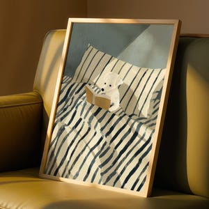 Op de afbeelding: Een ingelijste kunstdruk met een witte hond die een boek leest in bed. Het kunstwerk heeft een licht houten frame en toont een gezellige scène met een gestreept kussen en deken in tinten blauw en crème.